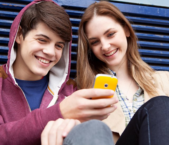 Eine junge Frau und ein junger Mann sitzen gegen eine Wand gelehnt und blicken lachend auf ein Smartphone. 