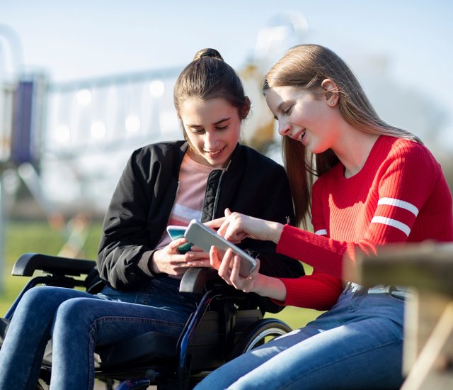 Zwei junge Frauen sitzen bei gutem Wetter draußen. Die linke Frau sitzt in einem Rollstuhl. Beide blicken auf ihr Smartphone und unterhalten sich darüber.