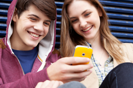 Eine junge Frau und ein junger Mann sitzen gegen eine Wand gelehnt und blicken lachend auf ein Smartphone.