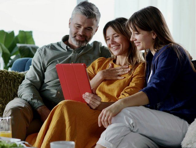 Ein Vater sitzt mit seinen zwei Töchtern auf dem Sofa. Sie schauen gemeinsam etwas auf einem Tablet an.